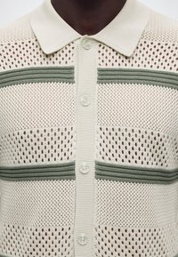 Κοντινό πλάνο εκρού πλεκτού πόλο πουκάμισου με πράσινες οριζόντιες ρίγες και κουμπιά μπροστά.