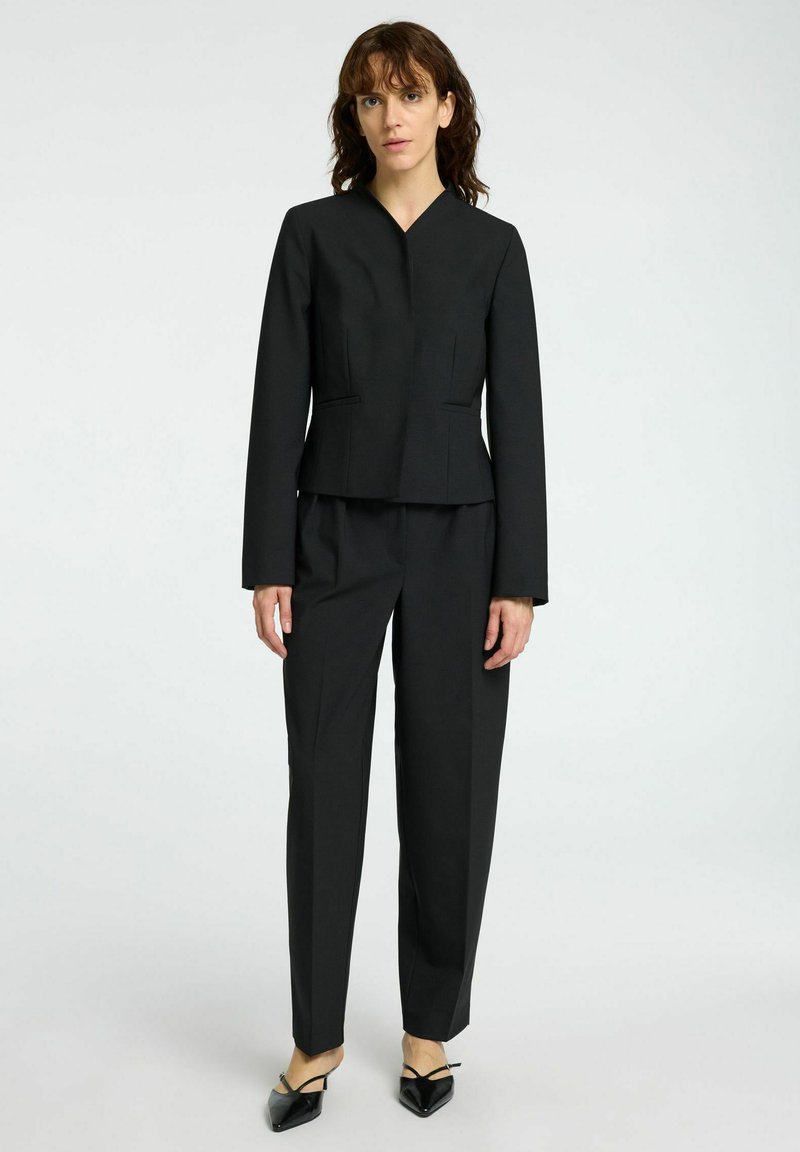 Schwarzes maßgeschneidertes Suit mit einer verkürzten Jacke, die einen V-Ausschnitt und Fronttaschen aufweist; kombiniert mit geraden Hosen und spitzen Schuhen.