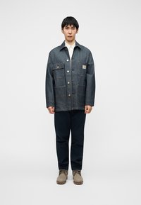 Τζιν μπουφάν με πολλαπλές τσέπες μπροστά, μεταλλικά κουμπιά και λογότυπο κέντημα. Φοριέται πάνω από ένα ανοιχτό πουκάμισο, συνδυασμένο με σκούρα μπλε παντελόνια και μπεζ παπούτσια.