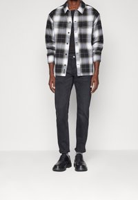 Svartvit rutad skjorta med knappdesign, i kombination med svarta jeans och svarta snörskor, på en neutral bakgrund.