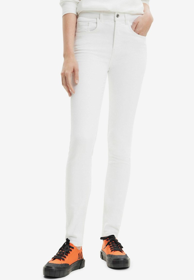Personne portant un jean blanc ajusté et des baskets basses orange avec semelles et lacets noirs, une main dans la poche.
