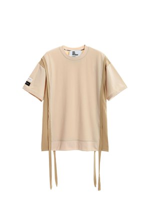 Joven de pie con las manos entrelazadas, vistiendo una camiseta beige extragrande, pantalones cortos a juego, calcetines blancos con estampado y botas de trabajo marrones sobre fondo blanco.