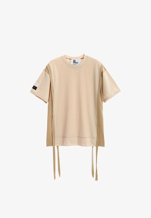 Joven de pie con las manos entrelazadas, vistiendo una camiseta beige extragrande, pantalones cortos a juego, calcetines blancos con estampado y botas de trabajo marrones sobre fondo blanco.