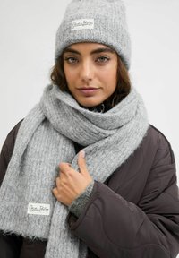 Naine, kes kannab halli kudumit mütsi ja sobivat salli, hoiab salli käes, riietatud tumedasse tikitud jopesse, lihtsa tausta taustal.