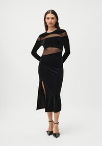 Melnas samta kleitas ar garām piedurknēm, kurām ir caurspīdīgas acs paneļi un sānu izgriezums. Dizains izceļ gludas tekstūras un pieguļošu formu.