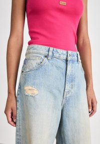 Femme portant un jean en denim bleu clair délavé et déchiré et un top sans manches rose vif avec un patch logo Hugo.