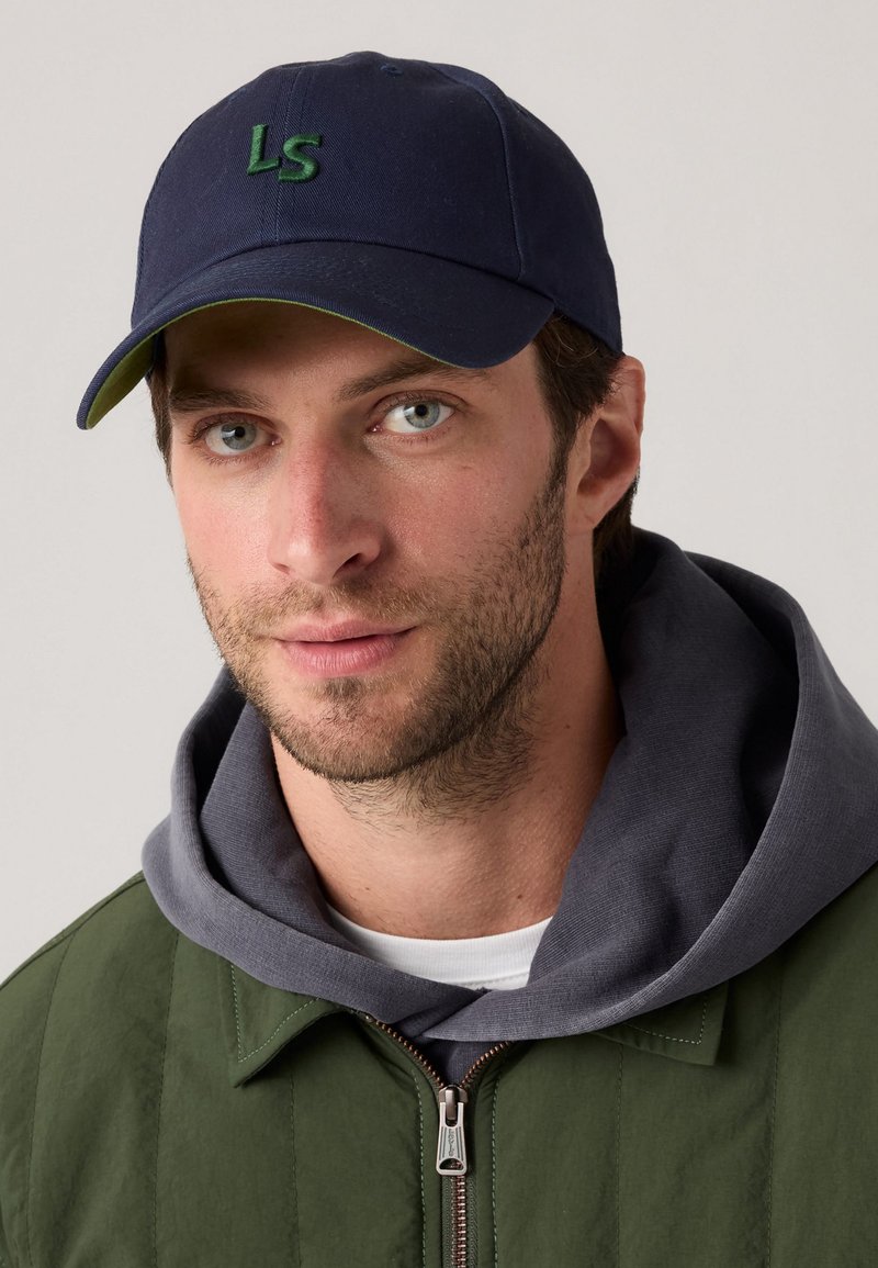 Casquette de baseball marine avec "LS" brodé en vert, associée à un sweat à capuche gris sous une veste zippée verte, ornée de rayures horizontales.