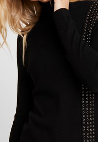 Blondynka w czarnym swetrze z metalowymi detalami na rękawach, z ręką blisko twarzy, na jednolitym tle.