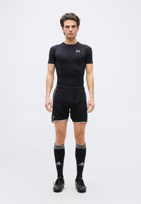 Νέος αθλητής με μαύρο συμπιεστικό μπλουζάκι, μαύρα αθλητικά σορτς, κάλτσες Adidas μέχρι το γόνατο και μαύρα παπούτσια ποδοσφαίρου, στέκεται μπροστά.