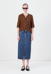 Brun strikket cardigan med korte ermer og knappelukking, kombinert med en denim maxiskjørt med høyt splitt foran og flate svarte sko.
