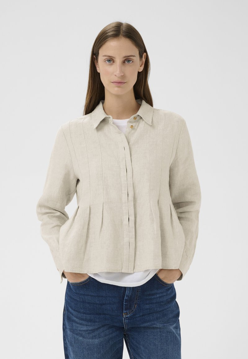 Femme aux longs cheveux bruns portant une veste en lin beige boutonnée sur une chemise blanche et un jean bleu, les mains dans les poches de la veste, fond neutre.