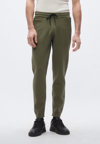 Pantalones de punto verde oliva con textura acanalada, cinturilla con cordón, bolsillos laterales y ajuste cónico, combinados con zapatillas negras.