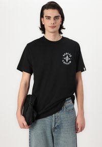 Svart bomulls T-skjorte med korte ermer, med et hvitt grafisk design på venstre bryst. Modellen holder en liten svart veske.
