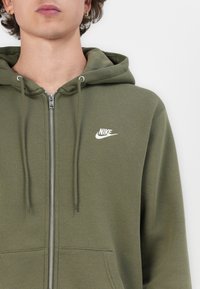 Sweatshirt verde-oliva com fecho, feito de um tecido suave, apresentando um bolso frontal, capuz com cordão e um pequeno logótipo branco da Nike no peito.