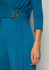 Teal innpakningsbluse med et gullfarget ovalt ring detaljer i midjen. Glatt stoff med en lett glans, tettsittende ermer og en strømlinjeformet silhuett.