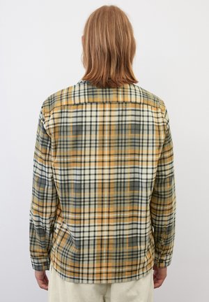 Personne aux cheveux châtain clair mi-longs portant une chemise à carreaux longue manche couleur moutarde, gris et blanc, de dos sur un fond uni.