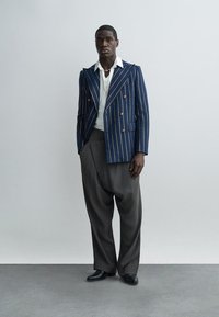 Mees seisab tavalise seina taustal, kandes mereväe sinist triibulist bleiserit, valget särki, lahtisi tumeda halli pükse ja musta jalatseid.