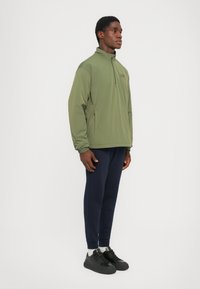 Olivenfarget hettegenser med glidelås og ståkrage, sidelommer og en glatt tekstur, kombinert med marineblå avsmalnende bukser og svarte joggesko.