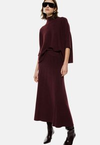 Maglione a costine bordeaux con scollatura alta abbinato a una gonna maxi a coste dello stesso colore. La modella indossa occhiali da sole oversize neri e stivali lucidi.