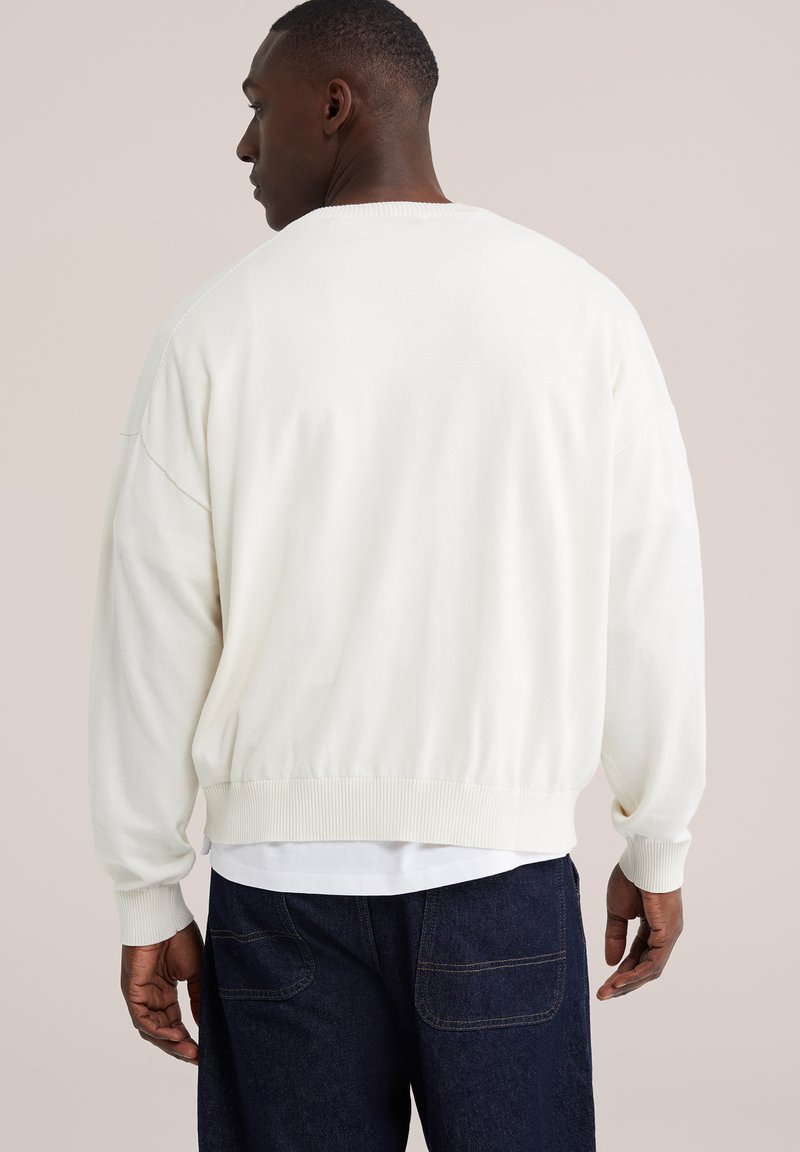 Homme vu de dos portant un pull blanc ample par-dessus une chemise blanche et un jean bleu foncé.