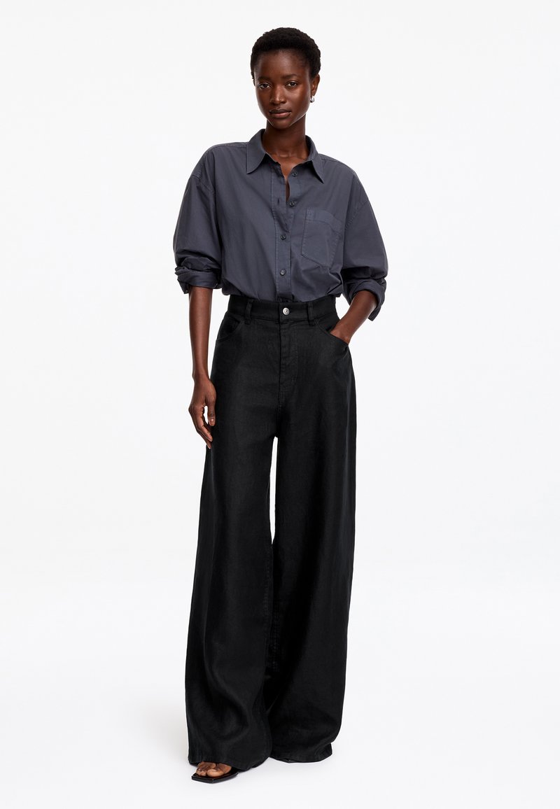 Personne debout, une main dans la poche, portant une chemise gris foncé à boutons et un pantalon noir à jambes larges, sur un fond blanc uni.