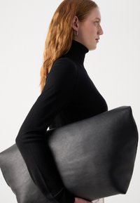 Černá kožená oversized tote taška držená proti černému přiléhavému roláku. Taška má hladký povrch, obdélníkový tvar a minimální kovové prvky.
