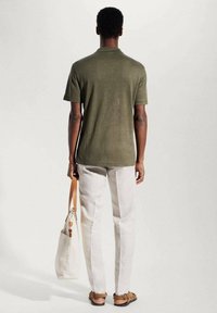 Mann in olivgrüner kurzärmeliger Hemd, hellbeigen Hosen und Sandalen, der eine weiße Baumwolltasche mit braunen Lederriemen trägt.