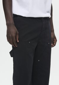 Zwarte cargobroek van een gestructureerde stof, met hoekige zakken, drukknoopsluitingen en een losse pasvorm. Vergezeld van een wit t-shirt.