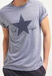 Šedé tričko s krátkým rukávem, které má tmavě modrou hvězdu a nápis "Chuck Taylor" napsaný kurzívou. Lehký materiál s volným střihem.