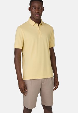 Helles gelbes Polo-Shirt mit Kragen und kurzen Ärmeln, kombiniert mit beigen Shorts. Verfügt über ein dezentes Logo auf der Brust. Feine Stofftextur.