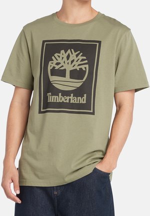 Mand iført en olivengrøn Timberland T-shirt med sort trælogo og tekst, kombineret med mørkeblå jeans.