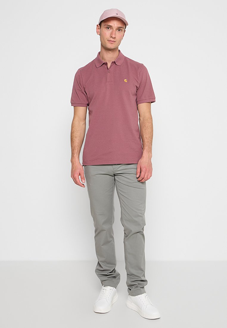 Mann steht mit einem mauvefarbigen Polohemd, passender Mütze, grauen Hosen und weißen Sneakers vor einem schlichten hellen Hintergrund.