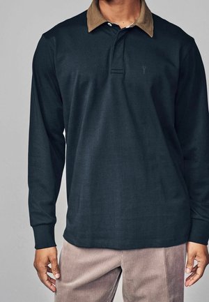 Mężczyzna ubrany w ciemną, długorękawową koszulkę polo z brązowym kołnierzem z sztruksu oraz beżowe spodnie sztruksowe, stojący na jednolitym, szarym tle.