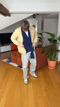 Beige jakke med glidelås, marineblå skjerf, hvit skjorte, lyseblå jeans og sølvfargede joggesko på et tregulv, med en plante og en sofa i bakgrunnen.