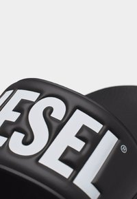 Μαύρο σανδάλι με φάσα και μεγάλο λευκό λογότυπο «DIESEL» στη φάσα, φωτογραφημένο από κοντά σε απλό φωτεινό φόντο.