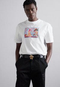 Camiseta blanca de manga corta con un estampado artístico y colorido en el frente. Combinada con pantalones negros y un cinturón con hebilla de león dorado.
