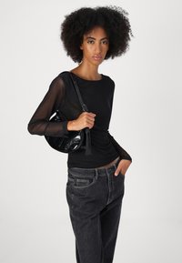 Black long-sleeve top with sheer sleeves, paired with high-waisted dark denim jeans. Model holding a small black handbag with glossy finish.