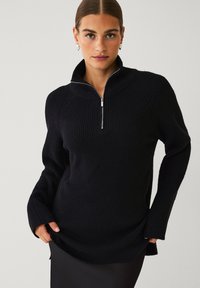 Sort strikket pullover med halv-zip krave, ribbet struktur, lange ærmer og sideslids. Stoflet ser varmt og behageligt ud.