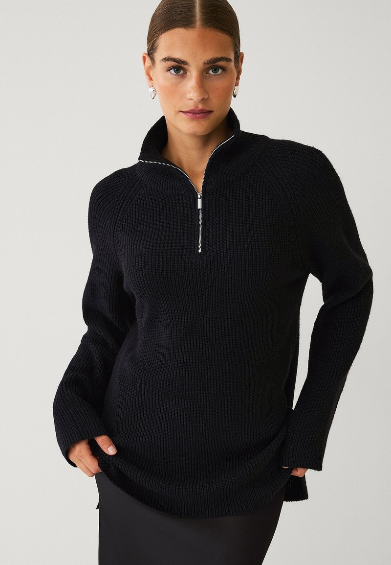 Sort strikket pullover med halv-zip krave, ribbet struktur, lange ærmer og sideslids. Stoflet ser varmt og behageligt ud.