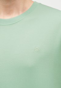 Gros plan d'une personne portant un t-shirt vert clair à col rond avec un petit logo brodé discret sur la poitrine gauche.