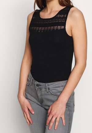 Frau trägt ärmelloses schwarzes Oberteil mit Spitzendetails am Ausschnitt und hellgraue Jeans, Hände auf den Oberschenkeln ruhend.