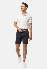 Polo shirt en coton blanc avec un logo, assorti à un short marine foncé et des baskets blanches. Ceinture avec des accents à motifs multicolores.