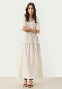 Long, cream-colored dress with short, embroidered sleeves, a drawstring waist, and decorative eyelet details near the hem. Black sandals worn.