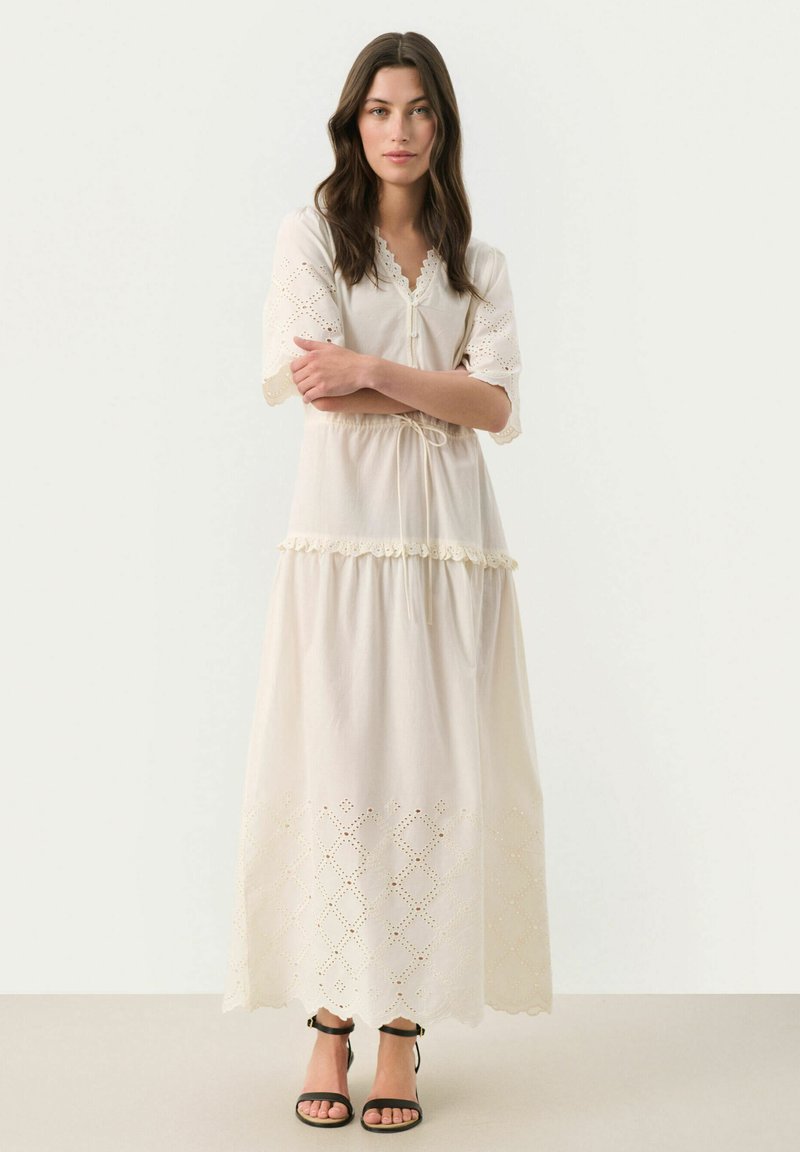 Long, cream-colored dress with short, embroidered sleeves, a drawstring waist, and decorative eyelet details near the hem. Black sandals worn.