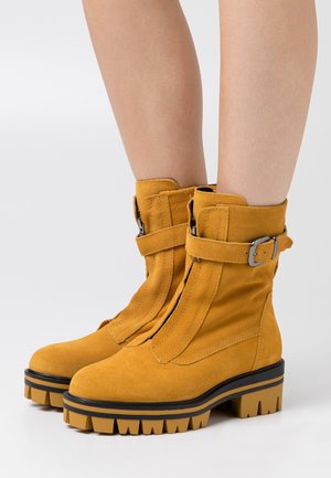 Des bottines en daim jaunes présentent une fermeture éclair à l'avant, une sangle ajustable, une semelle noire épaisse et une semelle en caoutchouc texturée.