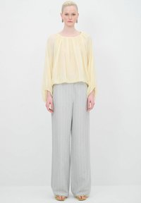 Gelbe Bluse mit gerafftem Ausschnitt und Ballonärmeln, kombiniert mit weit geschnittenen grauen Hosen mit Nadelstreifen. Model trägt Sandalen mit goldenen Akzenten.