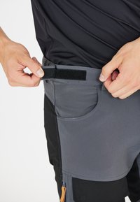 Grå shorts med justerbart midjeband i velcro, sidofickor och en kontrasterande svart panel. Texturen verkar vara slät och hållbar.