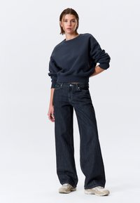 Sweat-shirt bleu marine à coupe décontractée, associé à un jean denim foncé à jambes larges et des baskets claires, avec des coutures simples et des accents légers.