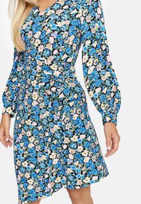 Osoba w kolorowej sukience w kwiaty z niebieskimi i różowymi motywami, z długimi rękawami i dopasowanym w pasie, stoi na jednolitym tle.