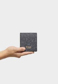 Cartera de cuero negro texturizado con un patrón en relieve, que presenta un logo dorado de "TOUS" en la parte frontal, sostenida en una mano. Forma cuadrada compacta.